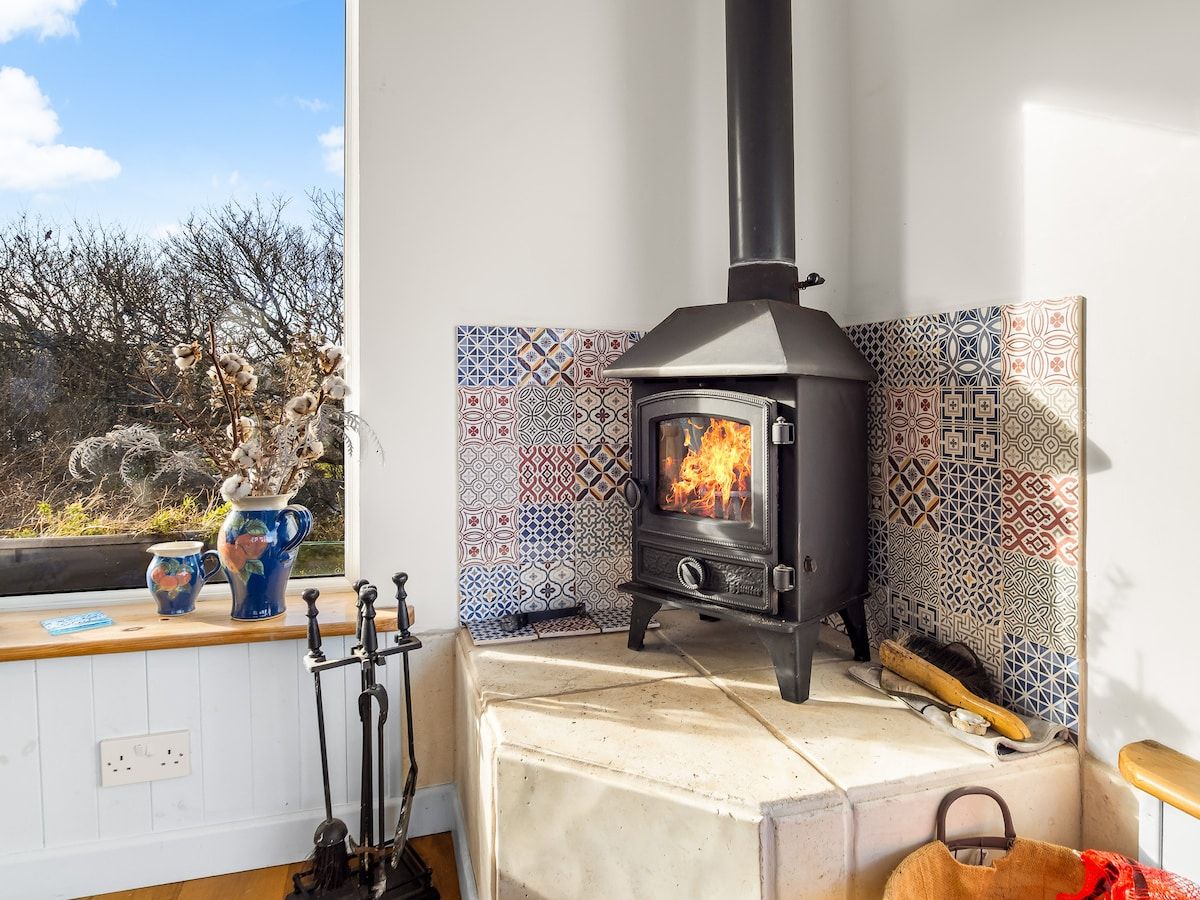 A room with a wood-burning stove, a window with a view of trees, and a tiled backsplash.