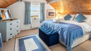 Une chambre avec un plafond en bois, un lit avec une literie bleue, une table de chevet avec une lampe, une commode avec des tiroirs, une fenêtre avec des rideaux et une vue sur la mer.