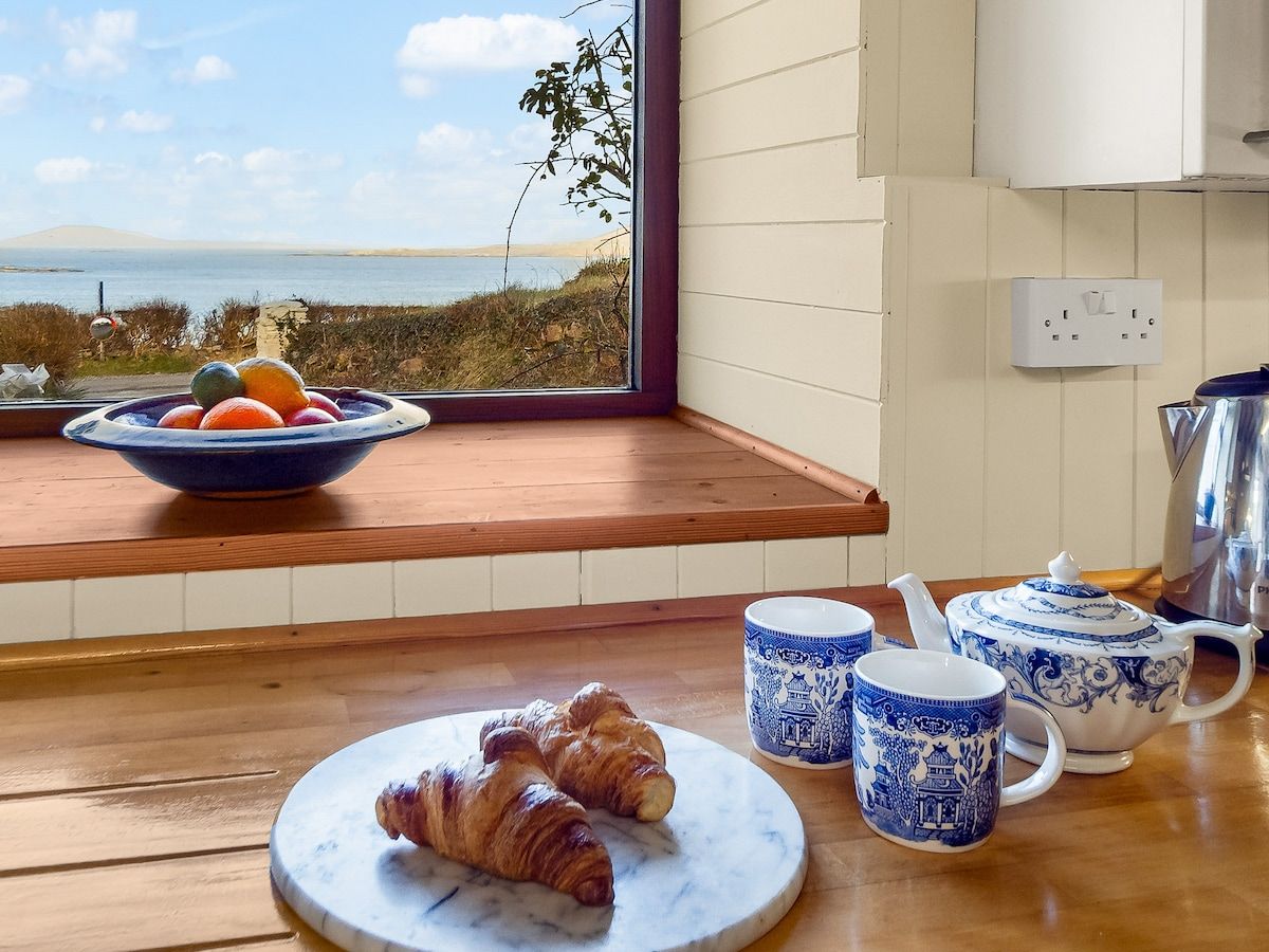 Une table en bois dressée pour le petit-déjeuner, avec des croissants, un saladier de fruits et des tasses bleues et blanches à motifs, le tout placé devant une fenêtre donnant sur la mer.