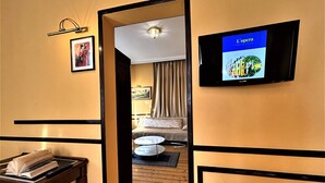 Een hotelkamer met een houten vloer, een bed met een beige deken, een klein rond tafeltje met een afstandsbediening en een boek, een aan de muur bevestigde televisie, en een ingelijst schilderij.