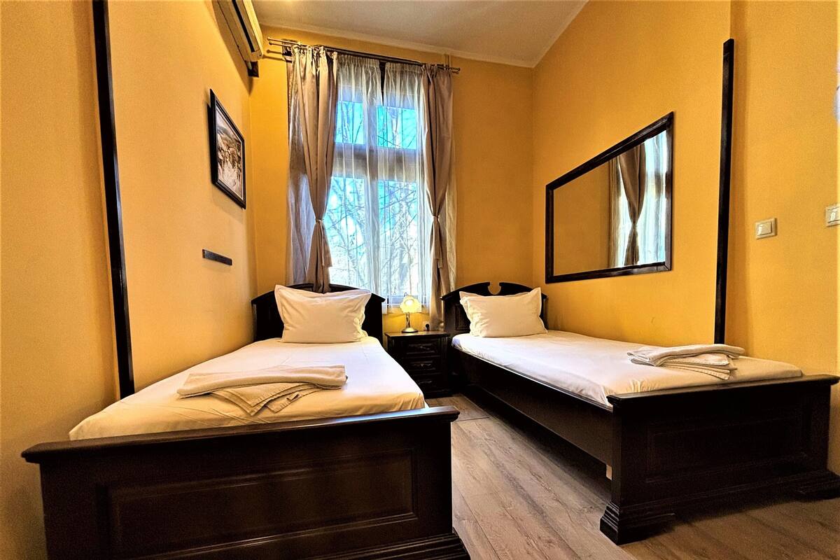A hotel room with two single beds, a bedside table with a lamp, a large mirror, and a window with curtains.