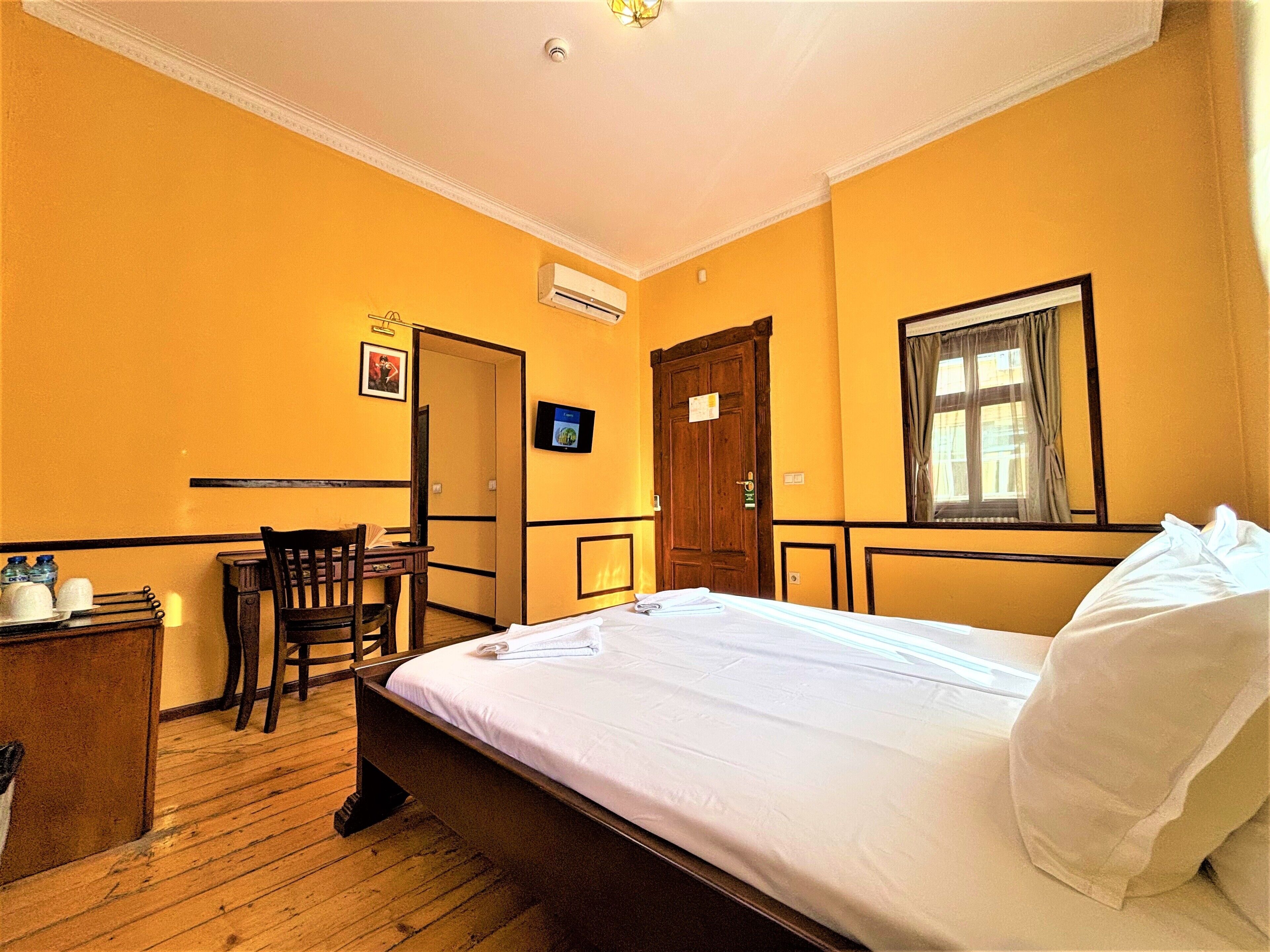 A hotel room with a wooden floor, a bed with white linens, a desk with a chair, a television, and a door.