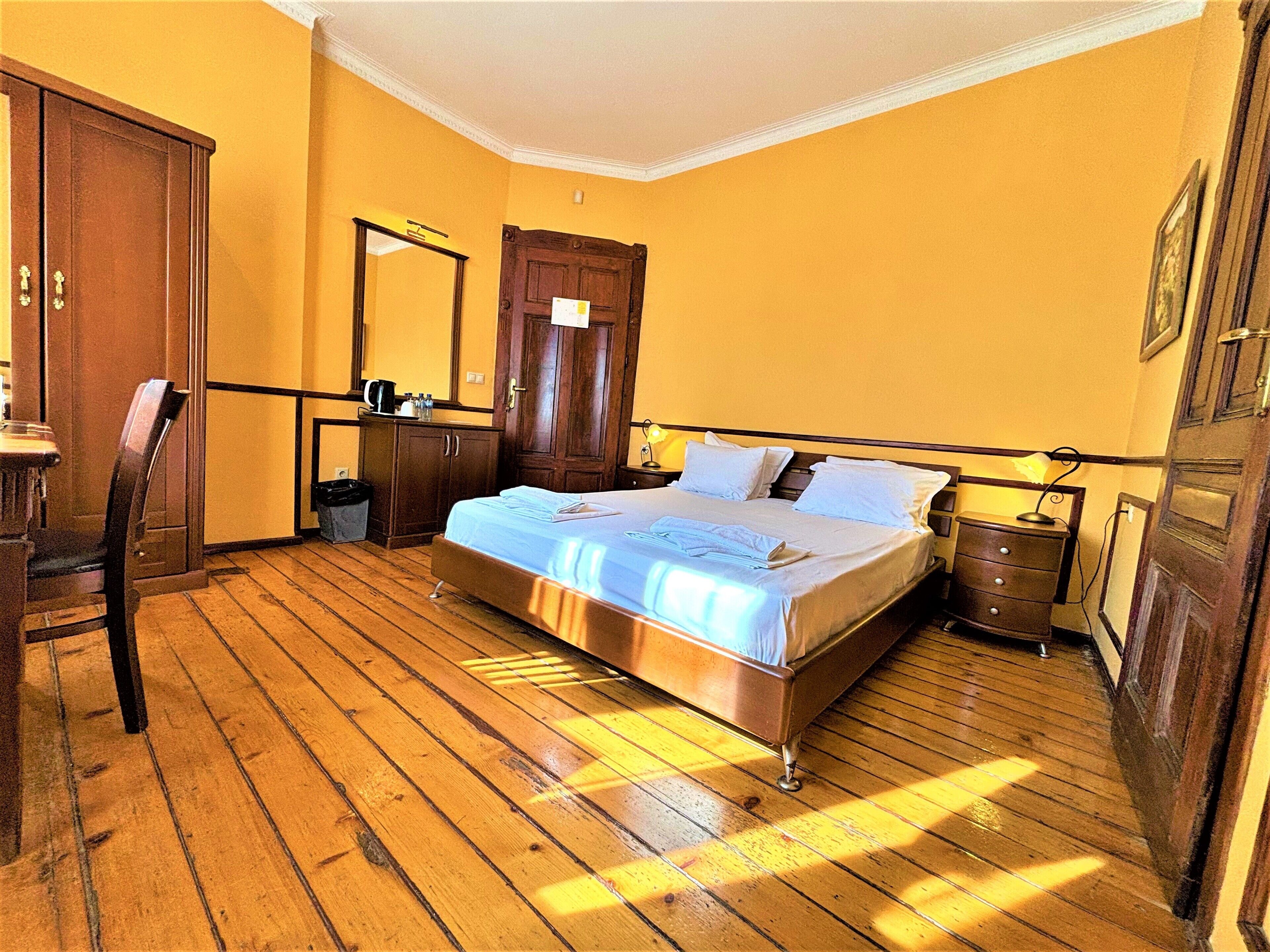 Un dormitorio con piso de madera, una cama con sábanas blancas, un armario de madera, un escritorio con una silla y un espejo en la pared.