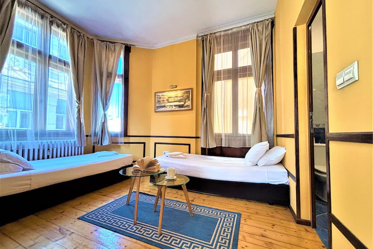 A hotel room with two beds, a small table, a window with curtains, a framed picture on the wall, and a wooden floor.