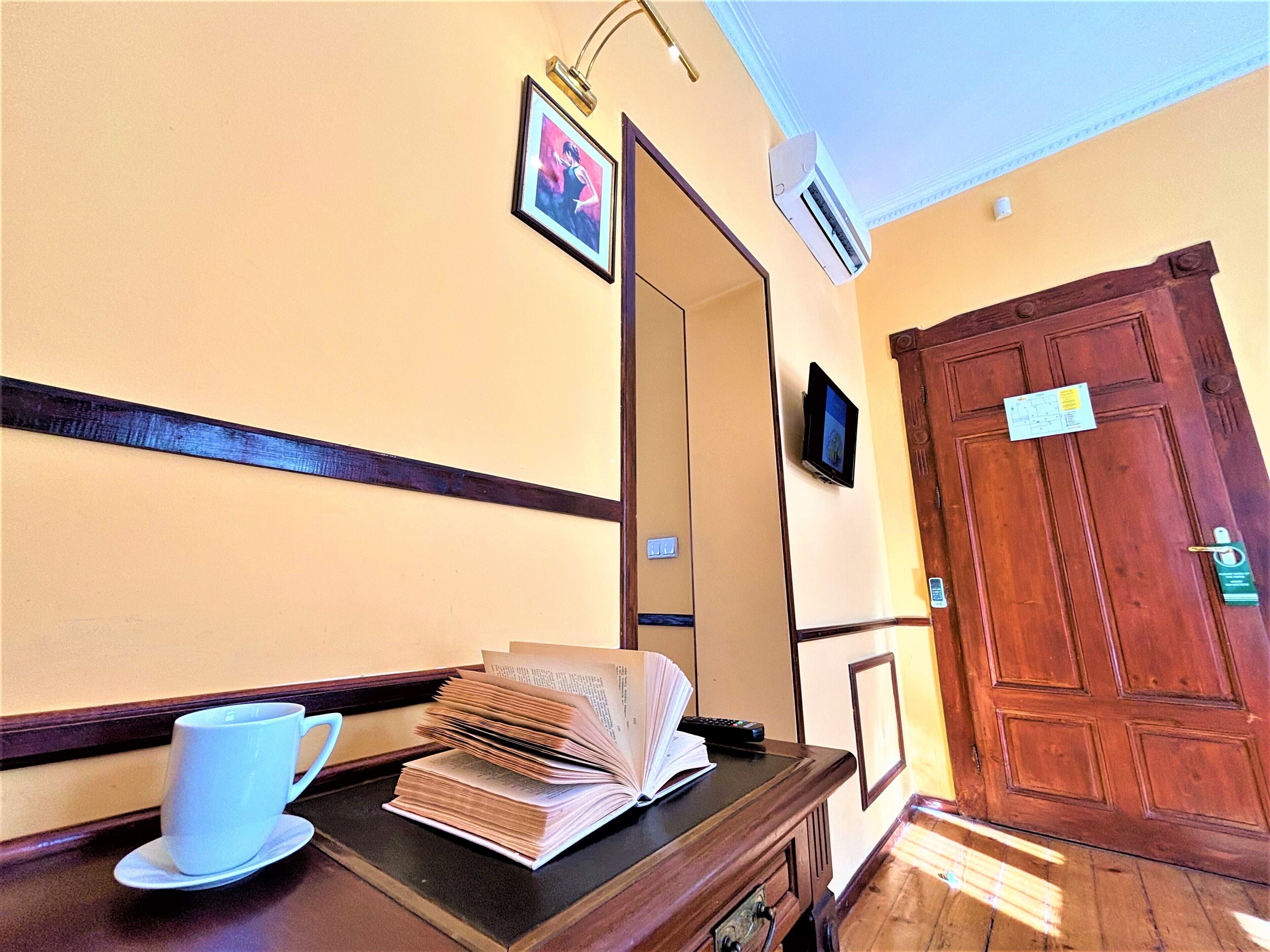 Uma área de recepção de hotel com balcão de madeira, uma xícara de café branca, uma pilha de livros, um quadro emoldurado e uma porta de madeira fechada.