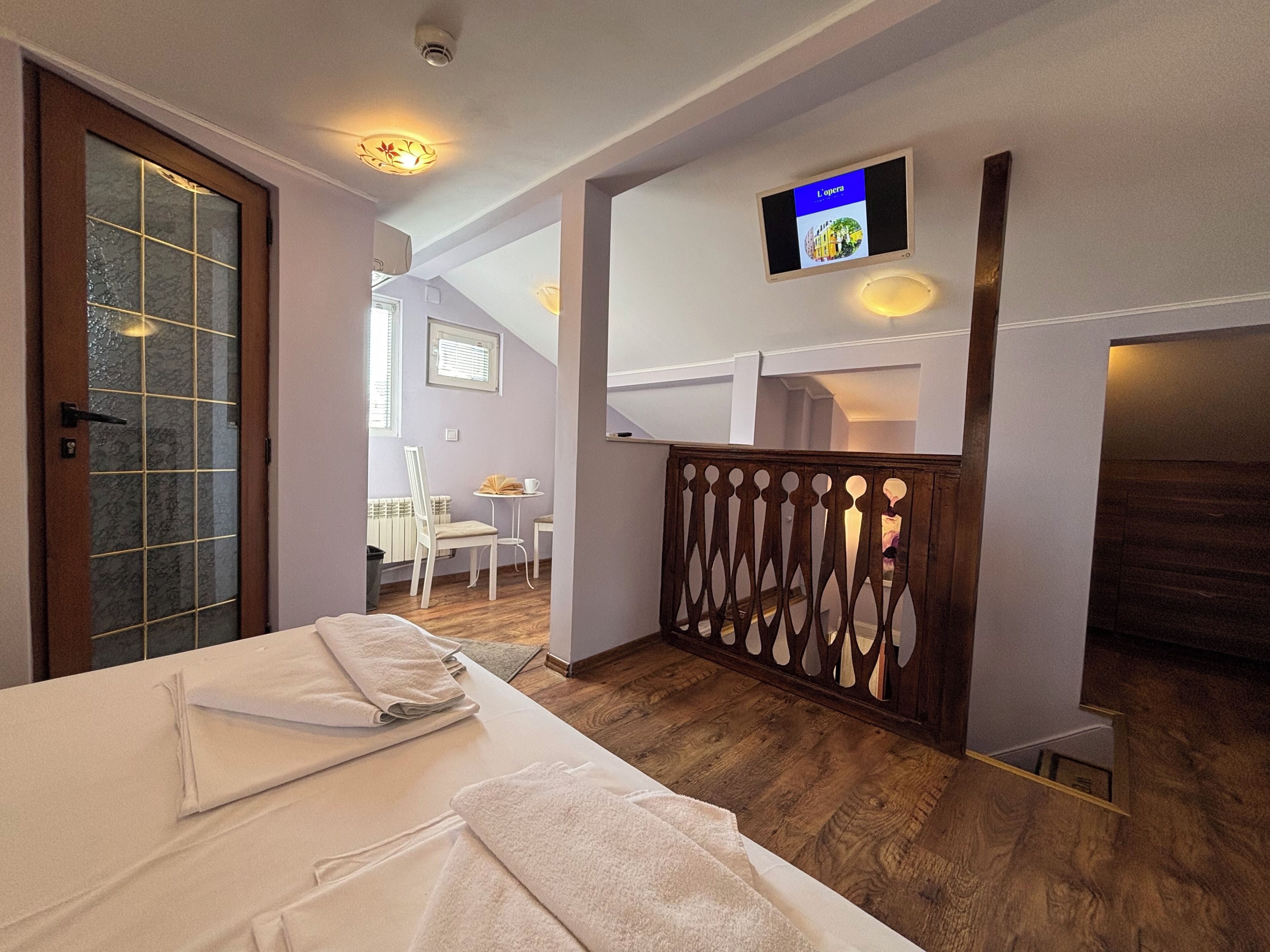 A hotel room with a bed, a small table with a chair, a TV mounted on the wall, and a wooden railing.