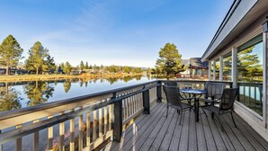 Une terrasse en bois avec une table et des chaises, donnant sur un lac paisible, entourée d’arbres et de bâtiments.