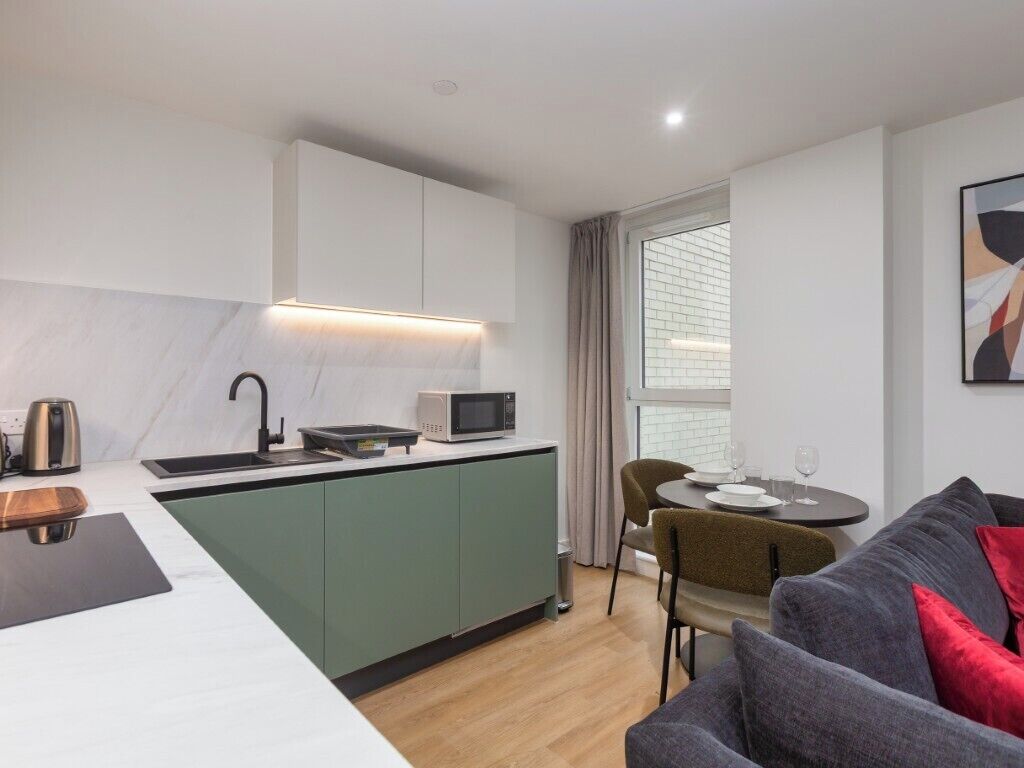 Cocina moderna con microondas integrado, hervidor y pileta. Hay un comedor con mesa y sillas, y un sofá gris con cojines rojos.