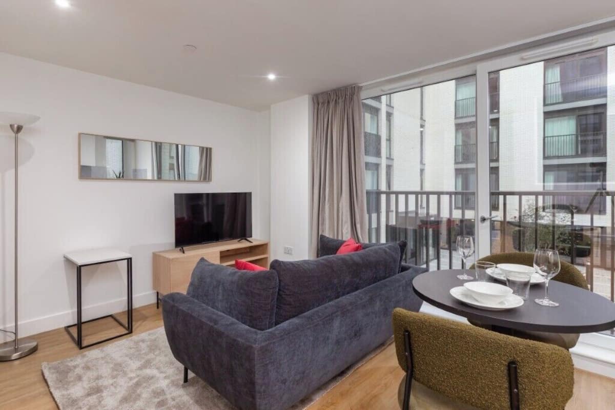 A modern living room with a sofa, a TV, a dining table with chairs, and a balcony with a view of buildings.