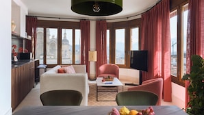 A modern living room with a sofa, armchairs, a dining table with fruit, a TV, and large windows with curtains.