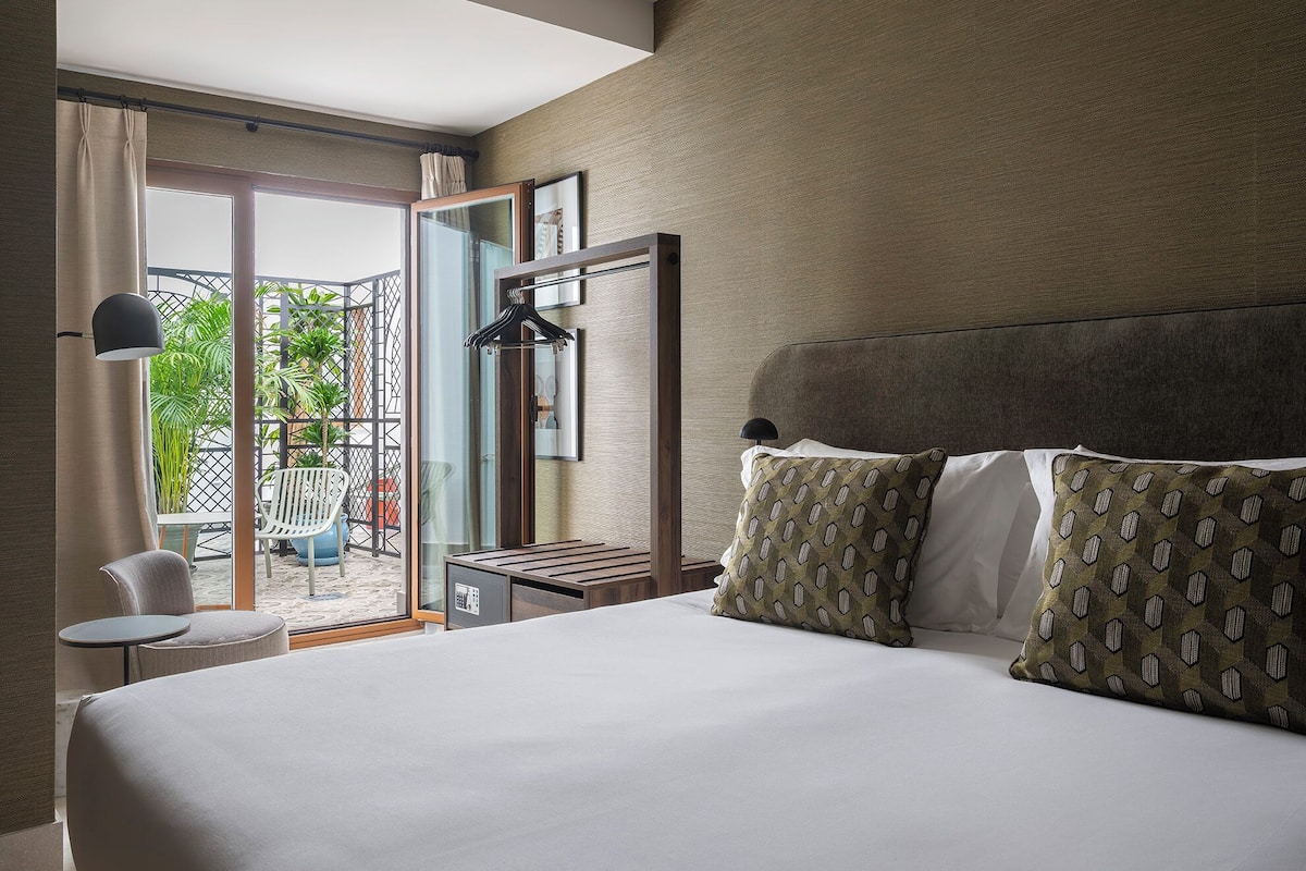 Habitación de hotel con cama, una mesita, una silla, una lámpara colgante y vistas al exterior.