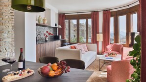 A modern living room with a sofa, a dining table with fruit and wine, a kitchen area, and large windows with a view.