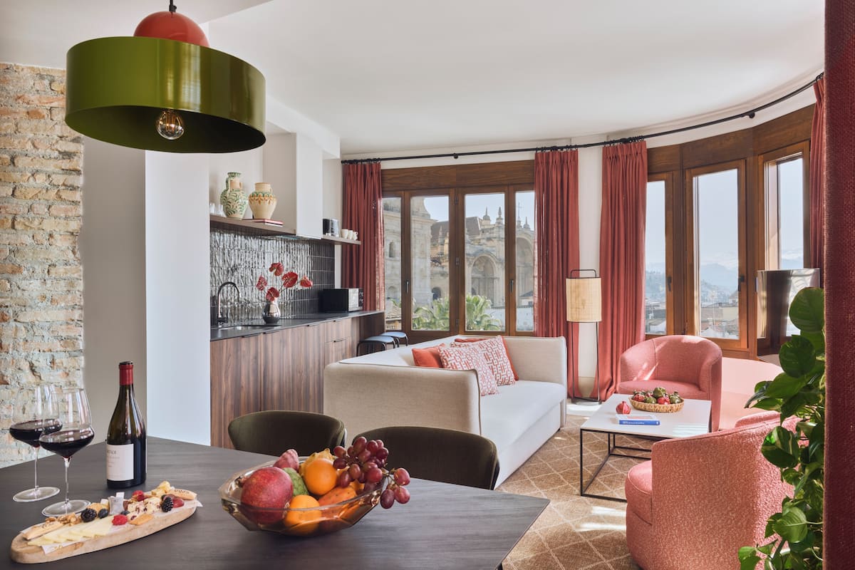 Una sala de estar moderna con sofá, una mesa de comedor con fruta y vino, una zona de cocina y ventanas grandes con vistas.