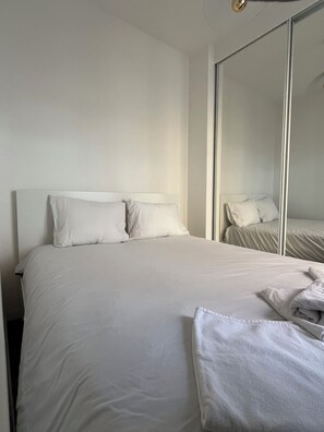 A neatly made bed with white linens and pillows, a bedside table with a lamp, and a large mirror reflecting the bed and a wardrobe. - Apartment Gare d'Austerlitz - Jardin des plantes (Paris)