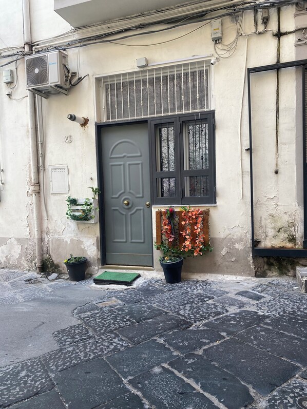 A building facade with a grey door, a window with metal bars, and a potted plant with orange flowers. - Queen Matthias Vacation Home (Napoli)