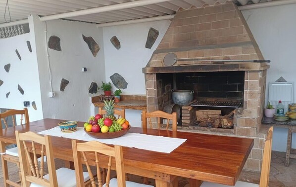 A rustic outdoor dining area with a brick oven, wooden table, chairs, and a bowl of fruit.