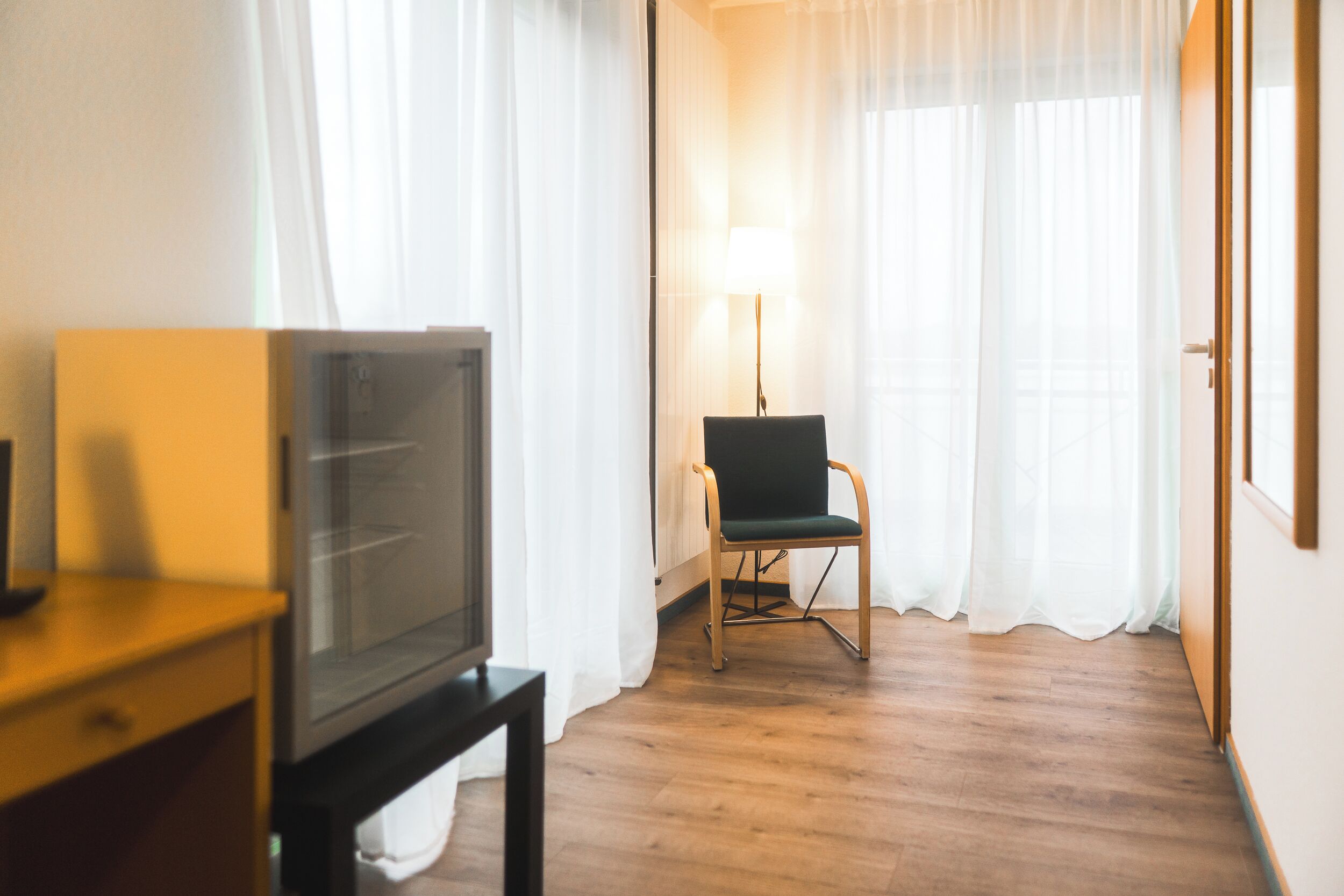 Een hotelkamer met een houten vloer, een televisie op een standaard, een stoel en een lamp.