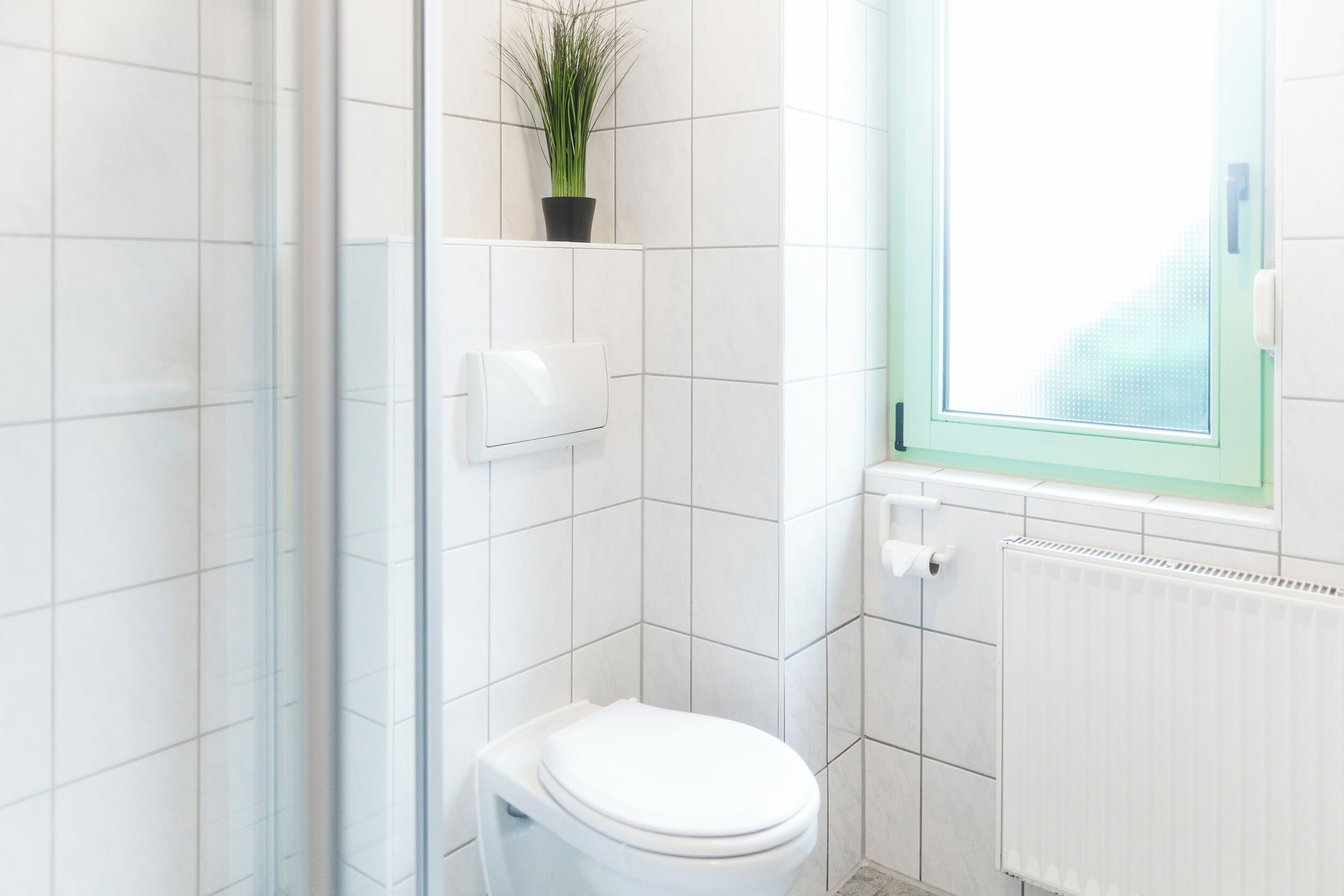 Un baño moderno con baldosas blancas, un inodoro, un secamanos, un radiador y una ventana con vistas.