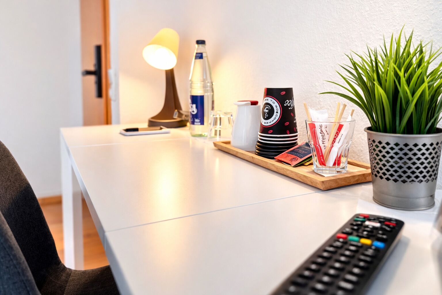 A clean, modern hotel room with a white desk, a lamp, a remote control, a plant, and a tray with various items including a water bottle, a coffee machine, and glasses.