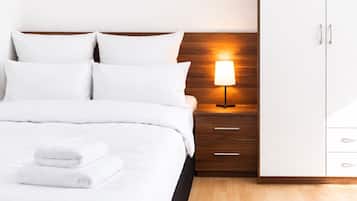 A neatly made bed with white linens and two folded towels, a bedside table with a lamp, and a wooden nightstand.