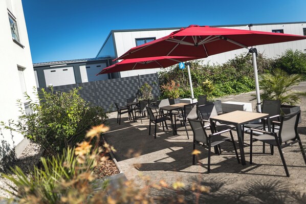 An outdoor patio with tables and chairs, a red umbrella, and a building in the background.