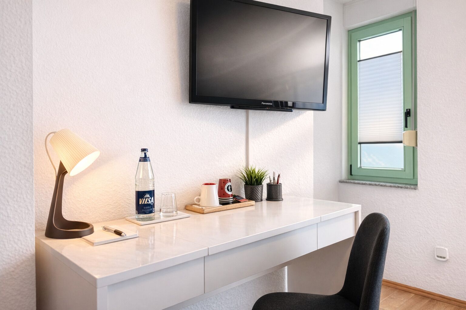 Una habitación de hotel moderna con un escritorio blanco, un televisor de pantalla plana, una lámpara de escritorio, una botella de agua, un vaso, una taza, una planta y una ventana con persianas.