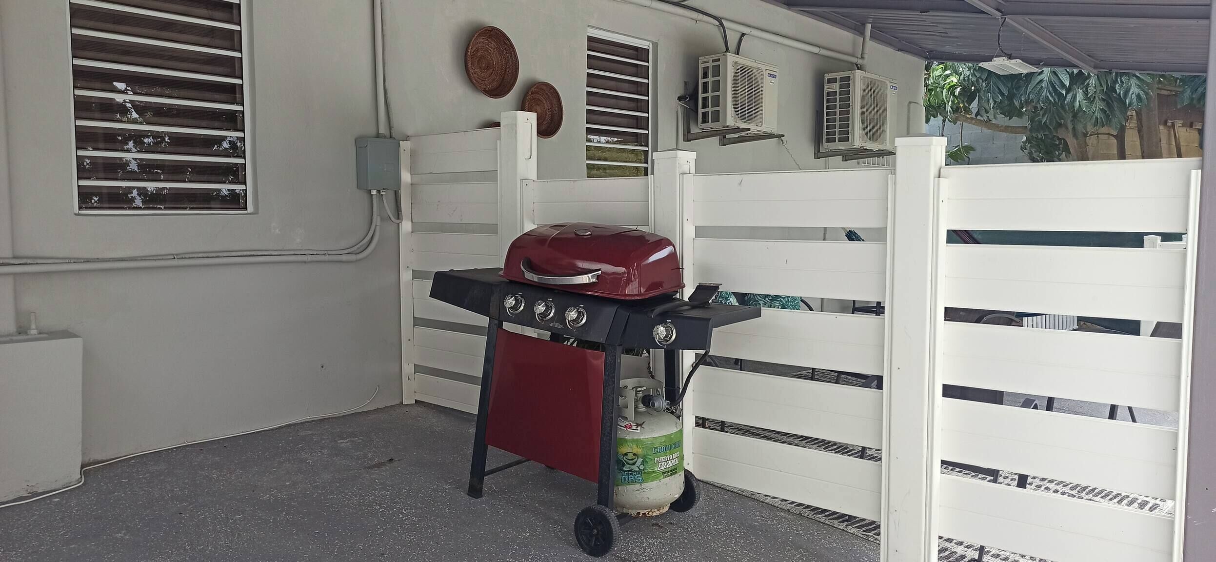Ein überdachter Außenbereich mit einem roten Grill, einer Gasflasche und einem weißen Zaun.