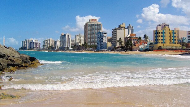 Un paisaje urbano costero con rascacielos, una playa arenosa y cielos azules despejados.