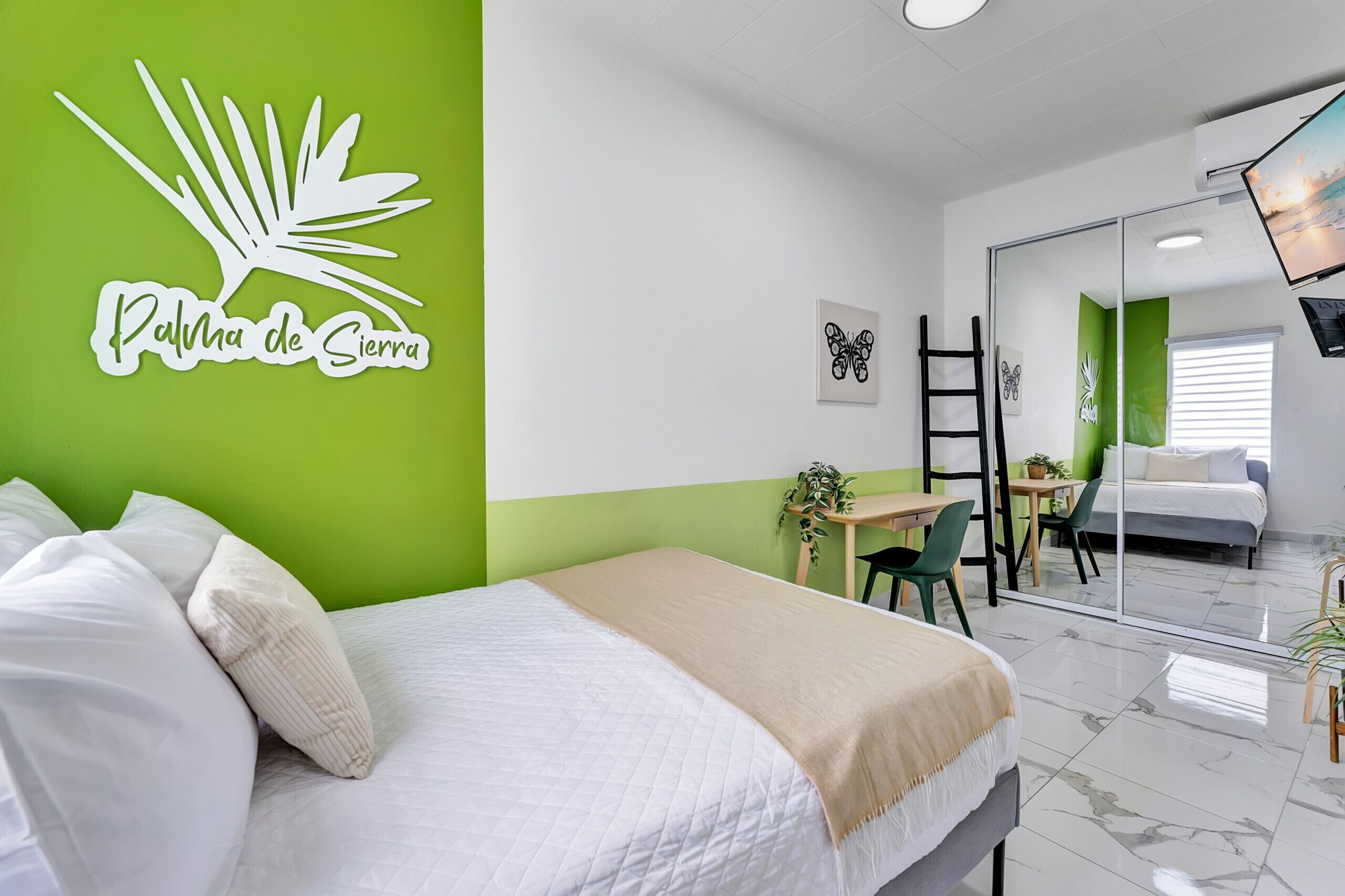 Une chambre d’hôtel avec un lit, un bureau et une chaise. Il y a un miroir au mur et une plante sur le bureau.