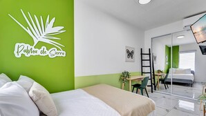 Ein Hotelzimmer mit einem Bett, einem Schreibtisch und einem Stuhl. An der Wand hängt ein Spiegel und auf dem Schreibtisch steht eine Pflanze.