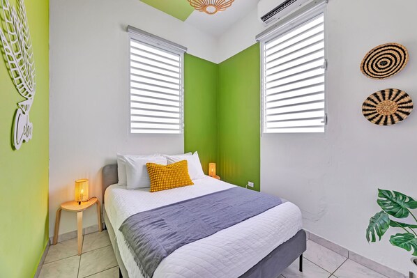 A bedroom with a bed, two bedside lamps, a wall-mounted air conditioner, and a green accent wall. - Centric, Spacious, Stylish Miramar Unit, Sleeps 4 (San Juan)