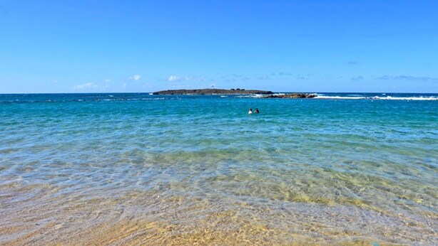 Ein Strand mit klarem, flachem Wasser, einem Felsen im Hintergrund und ein paar Schwimmern.