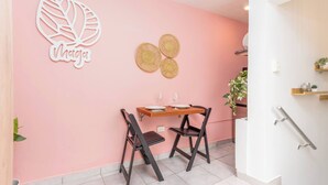 Una pared rosa con un adorno con forma de hoja y la palabra "Maga" debajo. A la derecha, una pequeña mesa de madera con dos sillas y una planta pequeña en maceta.