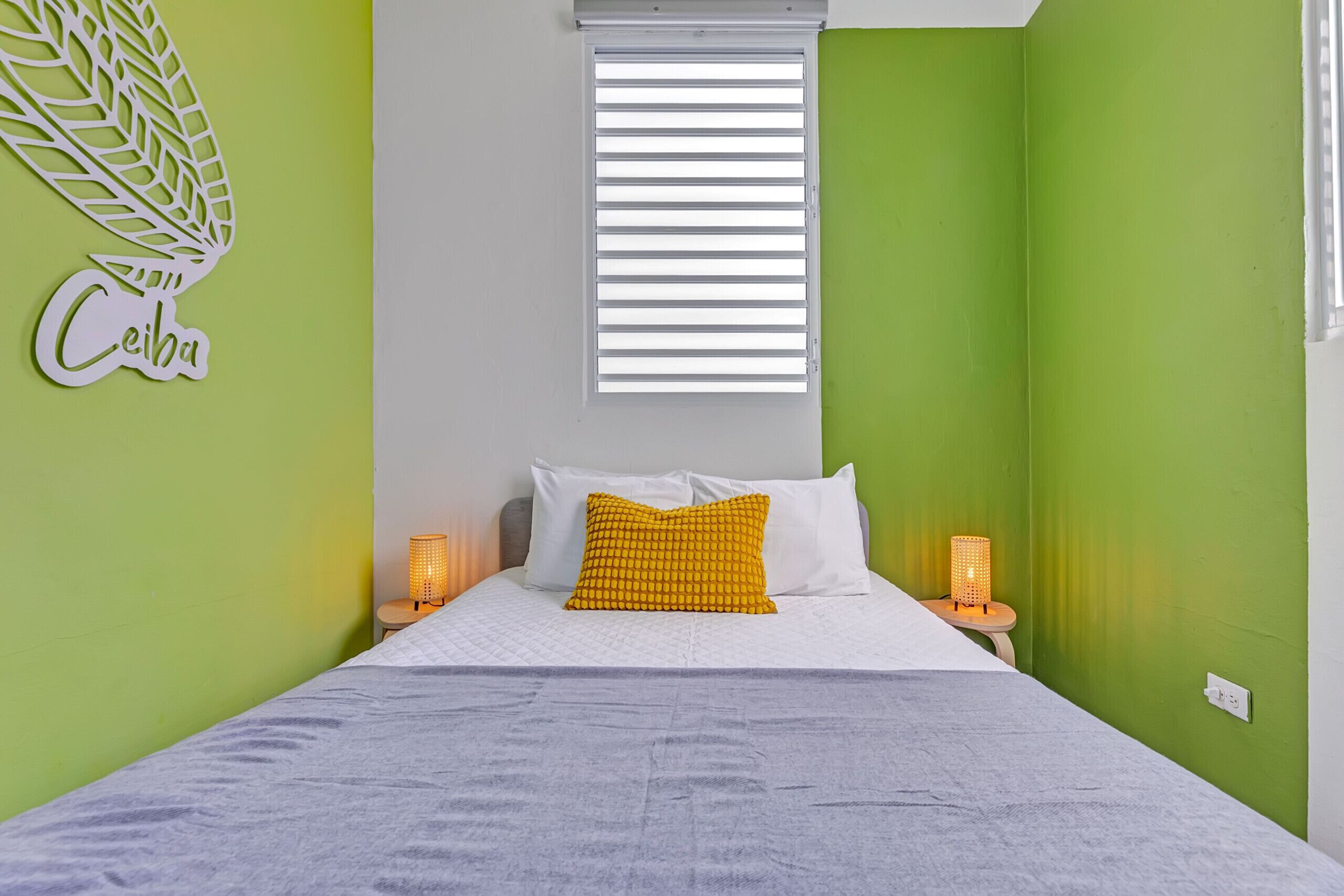 Une chambre à coucher avec un lit, deux lampes de chevet, une fenêtre avec des stores et un élément décoratif mural.