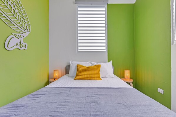 Ein Schlafzimmer mit einem Bett, zwei Nachttischlampen, einem Fenster mit Jalousien und einem Wandbild.