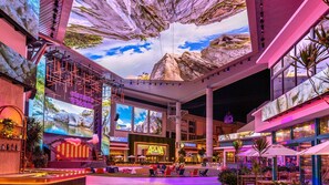 A spacious indoor atrium with a large digital screen displaying a mountainous landscape, a bar area with a neon sign, and a central seating area with outdoor views. - Bright, Centric and Cozy Studio In The Heart of Miramar (San Juan)