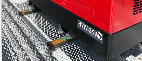 A red industrial generator with ventilation grilles, labeled 'HYW-25 M6,' on a metal mesh surface.