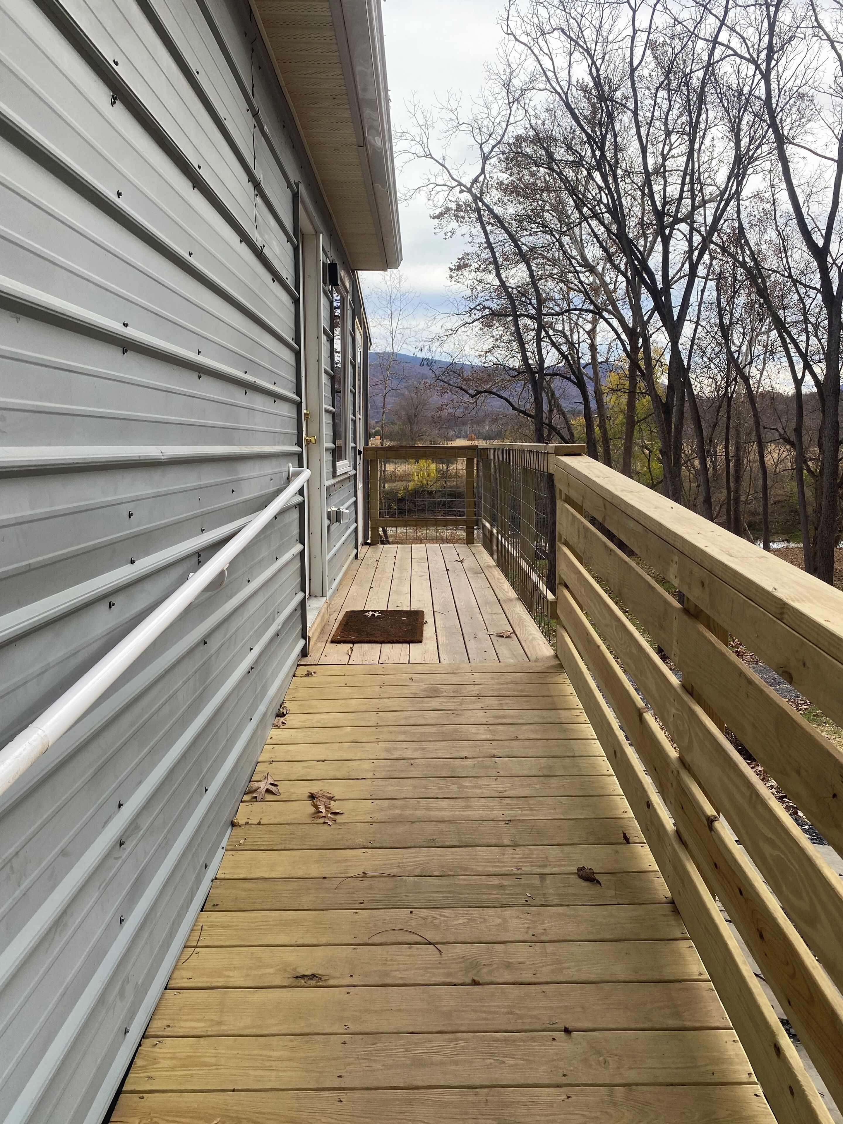 A wooden deck with a metal railing, leading to a covered area with a view of trees and hills.