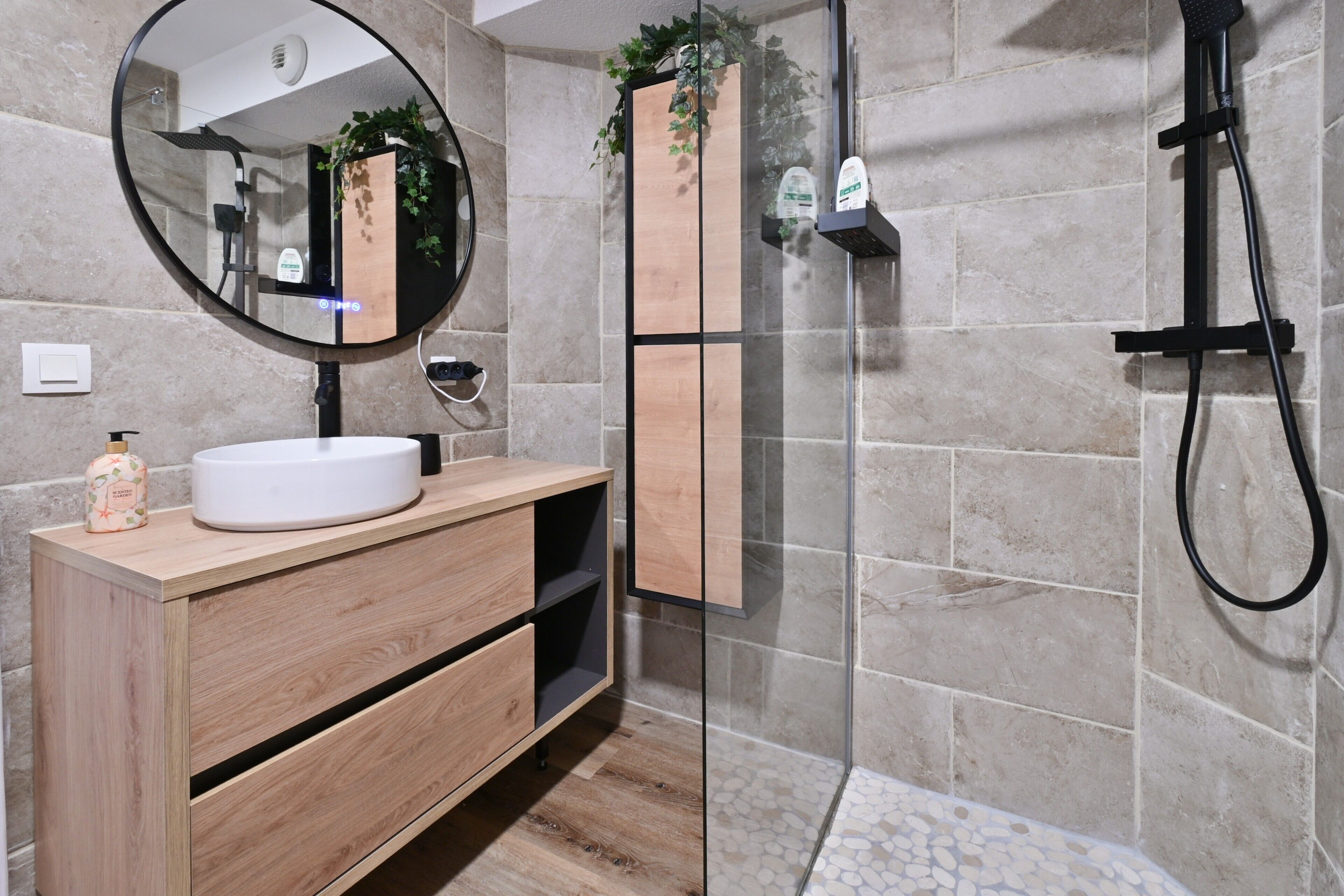 Een moderne badkamer met een ronde spiegel, een houten wastafelmeubel, een douchecabine van glas en betegelde muren.