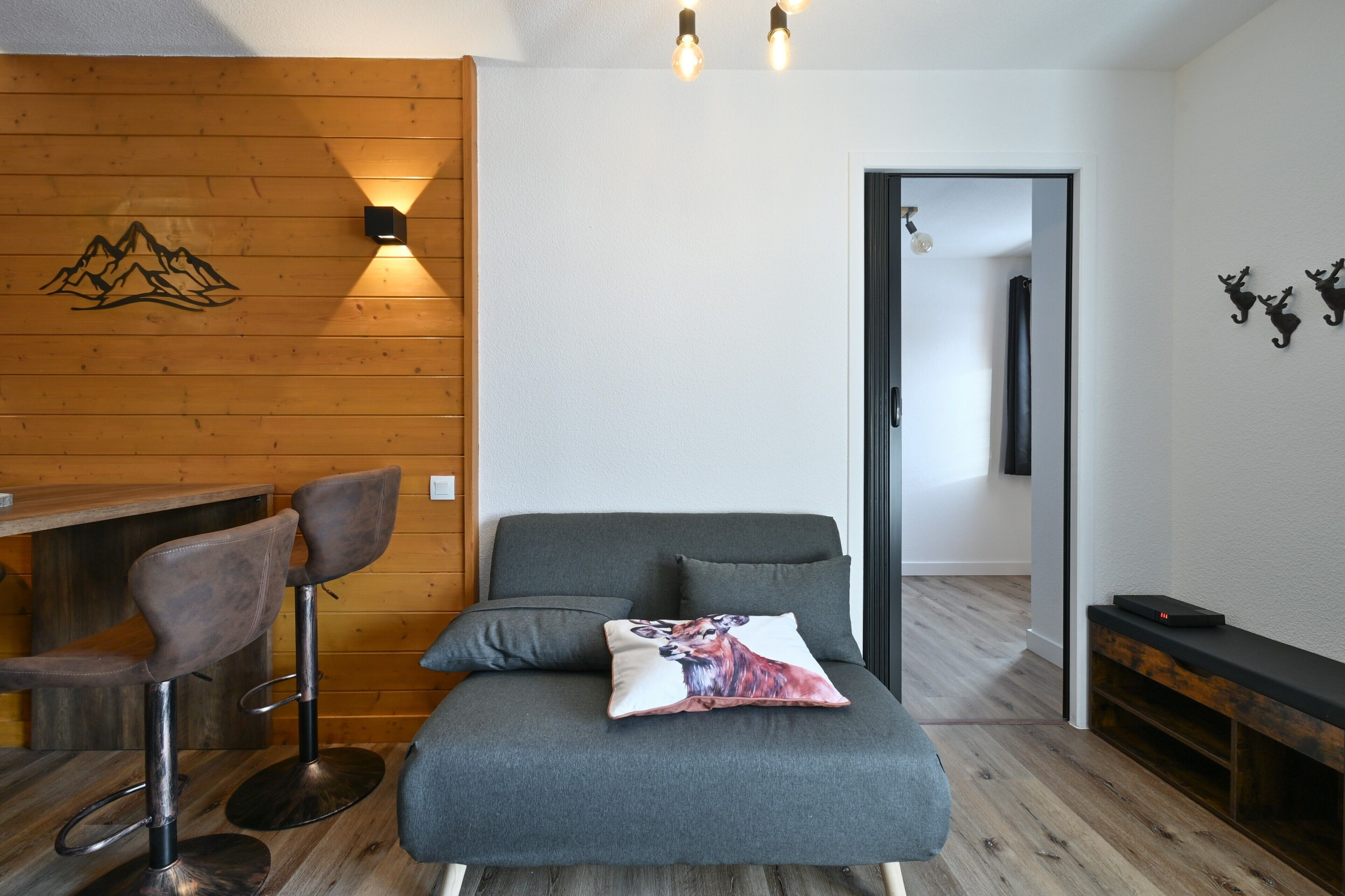 Une pièce avec un mur en bois, un canapé, deux tabourets de bar, un coussin orné d’un motif de renard et un banc.
