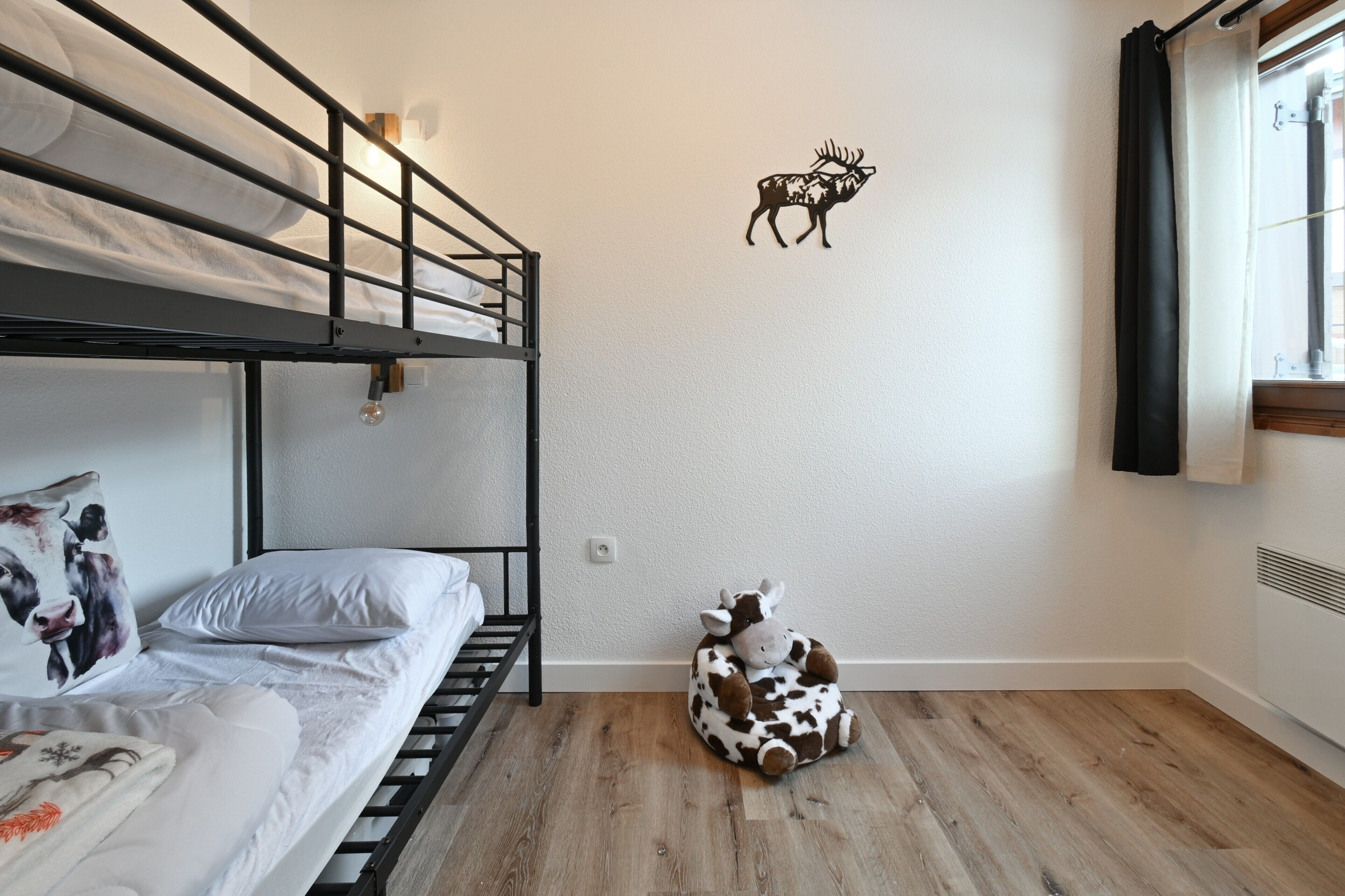 A bunk bed room with a cow plush toy, a wall decal of a deer, and a window with curtains.