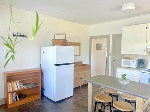 A compact kitchen with a white refrigerator, wooden cabinets, a small dining table with stools, and a bookshelf.