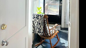 Una habitación con una mecedora de madera, una mesita con mantel estampado y un ventanal con vistas a árboles.