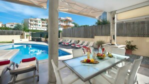 Zona junto a la piscina con sillas de descanso, una mesa con fruta y vistas a edificios al fondo.