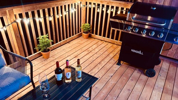 Una terraza de madera con una parrilla, plantas en macetas y una mesa con botellas y vasos.