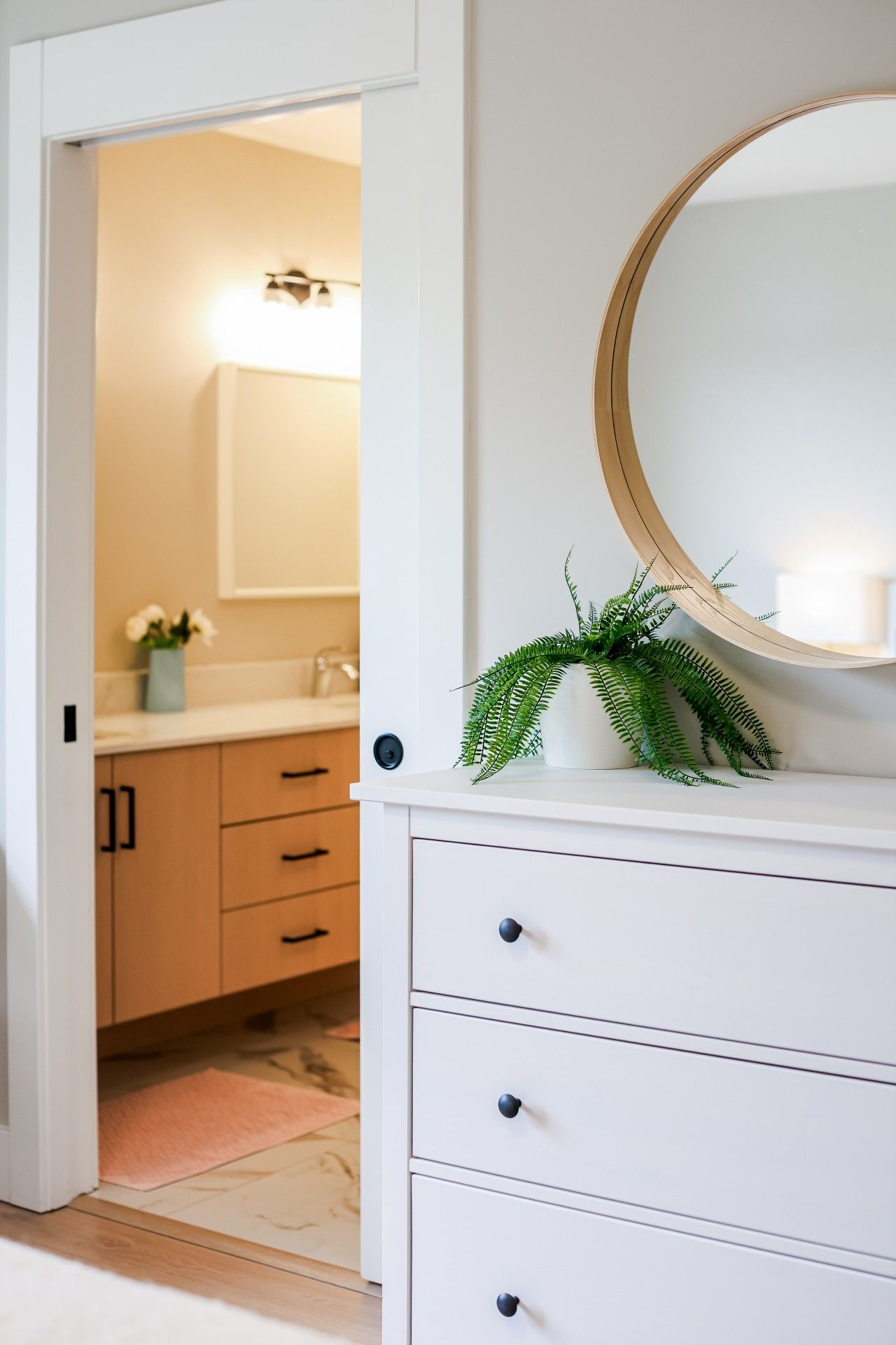 Une salle de bain avec un meuble-lavabo blanc, un miroir rond et une plante posée sur le plan de travail.