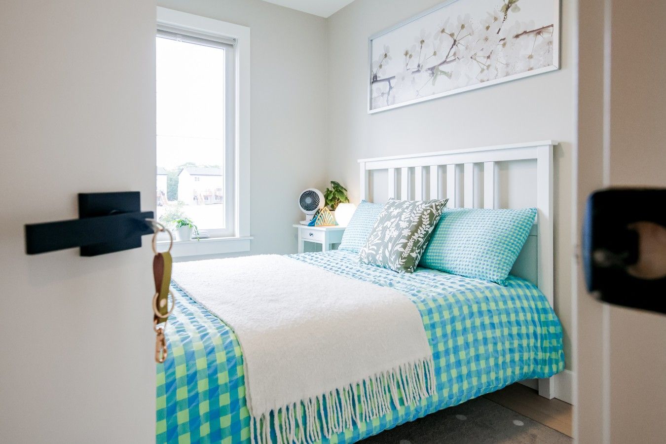 A bedroom with a bed, a window, a bedside table, a plant, and a wall-mounted artwork.