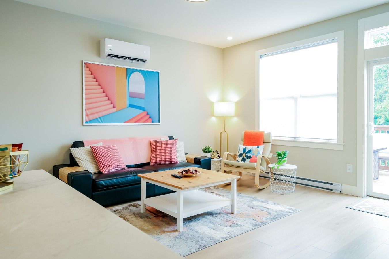 A modern living room with a sofa, coffee table, and a chair with a plant. There is a large window providing natural light and a painting on the wall.