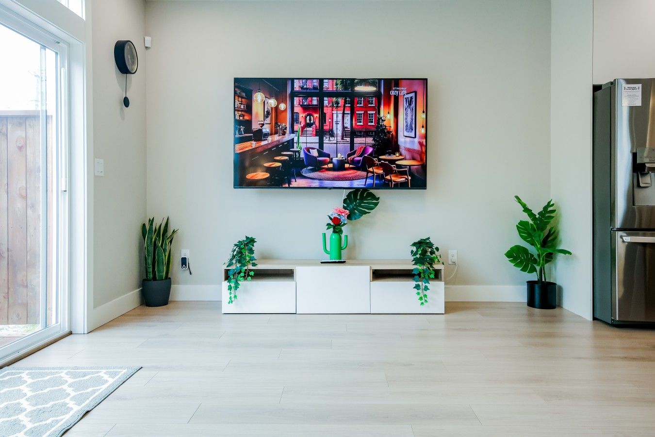 Un salon moderne doté d’un grand téléviseur à écran plat fixé au mur, d’un meuble de rangement blanc agrémenté de plantes et d’un réfrigérateur en acier inoxydable.