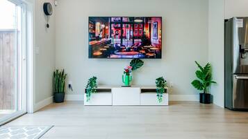 Un salon moderne doté d’un grand téléviseur à écran plat fixé au mur, d’un meuble de rangement blanc agrémenté de plantes et d’un réfrigérateur en acier inoxydable.